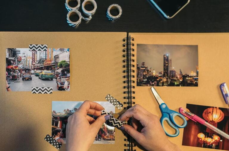 Image from above of the hands of an unrecognizable middle-aged woman picking up a piece of washi tape to make a handmade kraft travel album. Contains the word market in Thai. Image from above of the hands of an unrecognizable middle-aged woman picking up a piece of washi tape to make a handmade kraft travel album. Contains the word market in Thai.