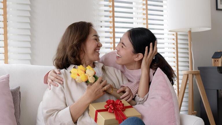 Attractive beautiful asian middle age mum sit with grown up daughter give gift box and flower in family moment celebrate mother day. Overjoy bonding cheerful kid embrace relationship with retired mom Attractive beautiful asian middle age mum sit with grown up daughter give gift box and flower in family moment celebrate mother day. Overjoy bonding cheerful kid embrace relationship with retired mom