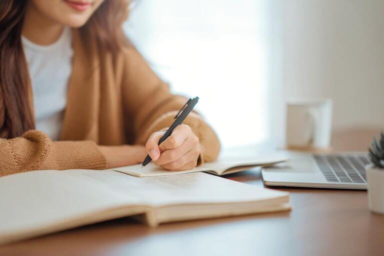 Closeup - Hand of woman writing in spiral notepad placed on wooden desktop with various items at home. Closeup - Hand of woman writing in spiral notepad placed on wooden desktop with various items at home.