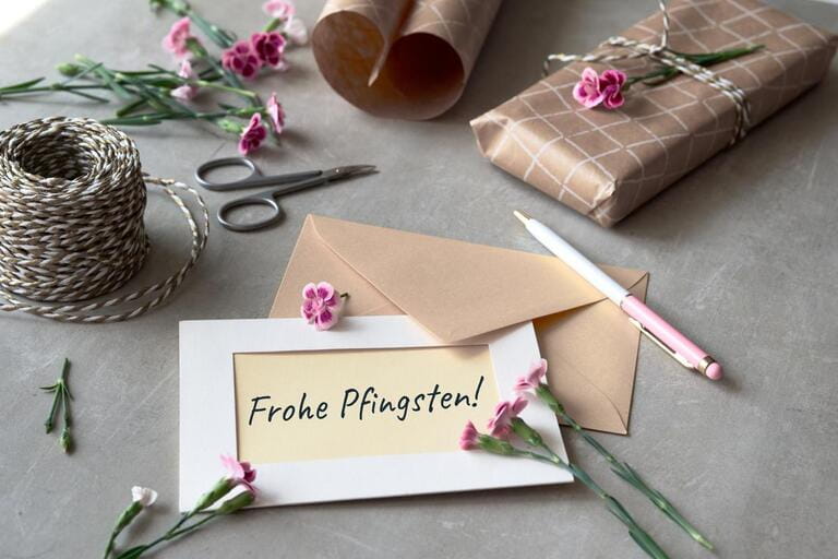 Text Frohe Phingsten means Happy Pentecost in German language. Envelope and greeting card with fresh carnation flowers. Wrapped gift, paper postcard, paper envelope, cord, and scissors on table. Text Frohe Phingsten means Happy Pentecost in German language. Envelope and greeting card with fresh carnation flowers. Wrapped gift, paper postcard, paper envelope, cord, and scissors on table.
