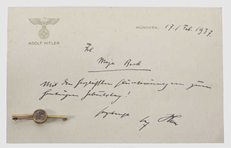 Maja Beck - a presentation brooch and birthday card from Adolf Hitler 1937,Gold brooch with pearl- and sapphire studded swastika in a circular,perforate filigreed bezel on a transverse bar with pin.Mark of fineness "585",length 48 mm,weight 2.6 g.The brooch pinned to a card with a raised national eagle in gold(darkened)above "Adolf Hitler" and the hand-written best wishes text "Frl.Maja Beck - Mit den herzlichsten Glückwünschen zum heutigen Geburtstag! Ergebenst Adolf Hitler"(Miss Maja Beck - With the most heartfelt best wishes on your birthday today!,Additional-Rights-Clearences-Not Available Maja Beck - a presentation brooch and birthday card from Adolf Hitler 1937,Gold brooch with pearl- and sapphire studded swastika in a circular,perforate filigreed bezel on a transverse bar with pin.Mark of fineness "585",length 48 mm,weight 2.6 g.The brooch pinned to a card with a raised national eagle in gold(darkened)above "Adolf Hitler" and the hand-written best wishes text "Frl.Maja Beck - Mit den herzlichsten Glückwünschen zum heutigen Geburtstag! Ergebenst Adolf Hitler"(Miss Maja Beck - With the most heartfelt best wishes on your birthday today!,Additional-Rights-Clearences-Not Available